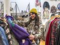Junge Frau lächelt, als sie in einem Secondhand Laden ein farbenfrohes Regal mit gebrauchter Kleidung durchsucht und nac